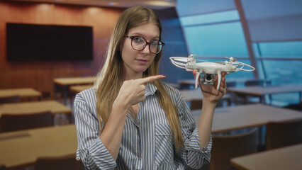 Young blonde woman wearing eyeglasses holding a white quadcopter drone in building; innovation curiosity.