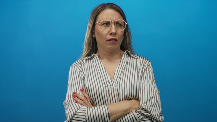 Woman with blonde hair wearing glasses and striped blouse frowns and crosses arms in blue studio;...