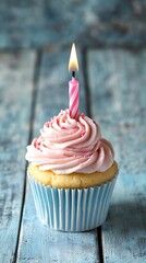 Cupcake with pink frosting, sprinkles, and burning birthday candle on blue wooden surface