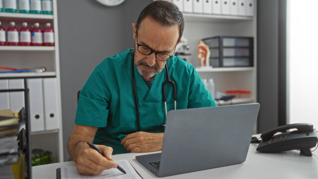 Mature hispanic man working in a clinic setting with medical equipment and documents, wearing a green uniform and glasses while using a laptop and writing notes at the desk