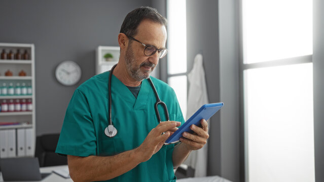 Mature hispanic male doctor in scrubs using tablet in well-lit hospital room, with stethoscope around neck, focused on digital healthcare tasks in modern medical setting