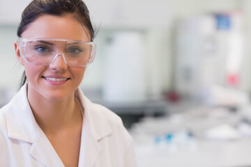 Smiling female scientist wearing lab coat, safety goggles at lab bench with pipettes, copy space