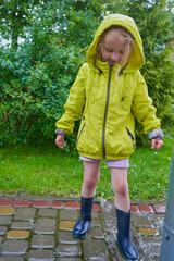 A little girl is playing in a puddle, happy little girl in a yellow jacket and rubber boots playing...