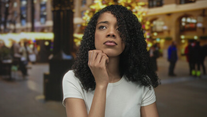 African american woman touches chin on city street beside illuminated christmas tree lights;...