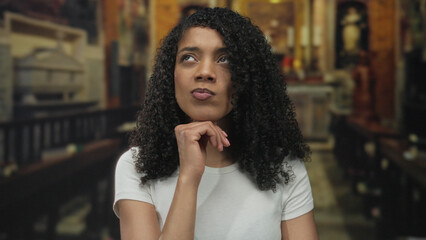 African american woman with hand on chin seated on wooden pew in church sanctuary; contemplation...