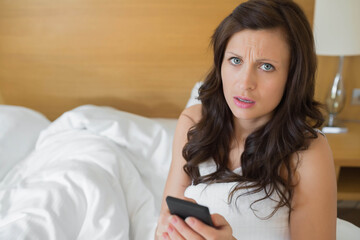 Woman holding smartphone sitting on bed with headboard looking concerned by lamp on bedside table