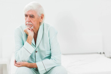 Senior man in light green pajamas sitting on white bed looking right in bedroom, copy space