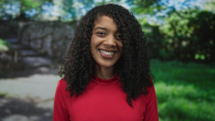 Woman smiling in red shirt in forest beside stone wall under bright sunlight looking to side;...