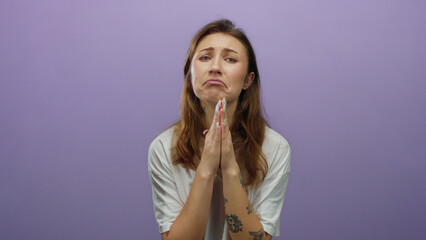 Woman with tattoos in a white shirt pleading with hands together in front of a purple background,...