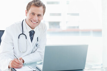 Male adult doctor writing on clipboard at glass desk with laptop and stethoscope, copy space