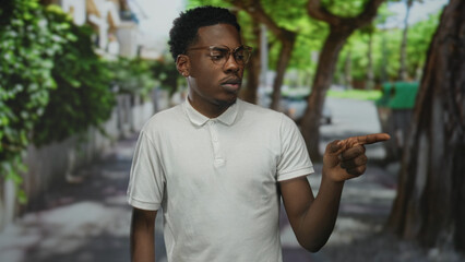 Young man wearing glasses and white polo shirt pointing finger to object on a sunlit city street; confidence.