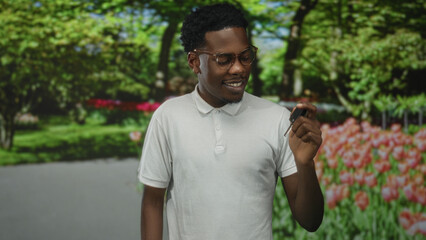 Fototapeta premium Man holding key remote with fingers and smiling in green park path lined by tulips and trees; confidence.