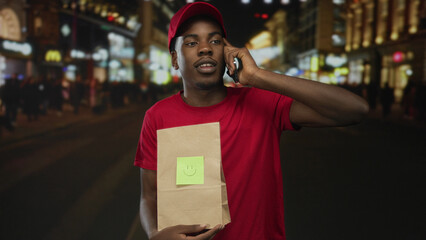Man holding delivery bag while talking on smartphone to ear on crowded busy city street at night;...