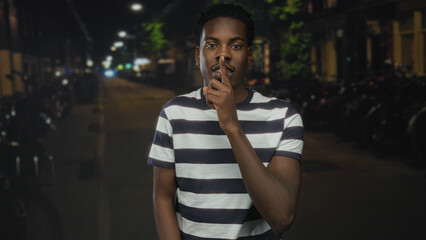 Man wearing striped shirt with finger to lips for silence on city street at night; silence secrecy...