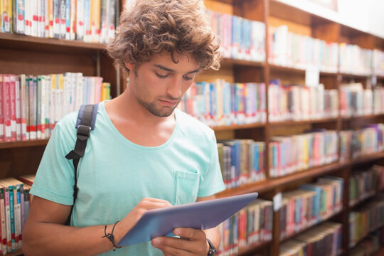 Man standing between wooden bookshelves in library aisle scanning tablet screen wearing backpack - Powered by Adobe