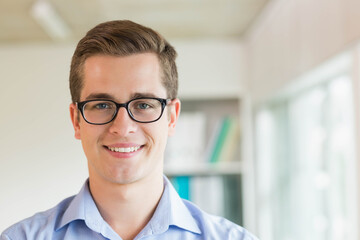 Male standing in office wearing glasses, light blue shirt by shelf with binders and window blinds