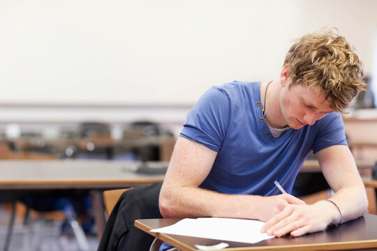 Male student leaning over wooden desk writing on paper in school classroom with pen, copy space