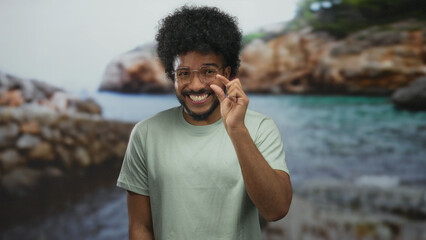 Smiling man with curly hair in glasses making small gesture at seaside with rocks and ocean in background.