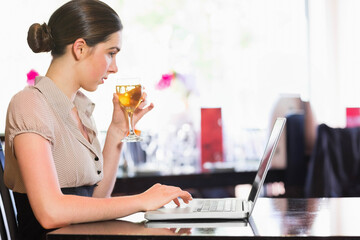 Woman holding stemmed glass and typing on silver laptop at cafe table with flowers, copy space