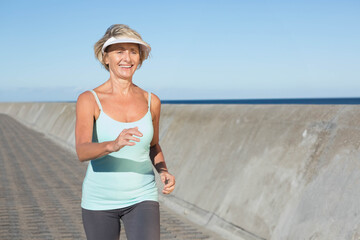 Senior woman jogging along promenade by sea wall wearing light blue tank top with sun visor