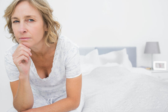 Senior woman sitting on bed edge, resting chin, gazing around bedroom with lamp, copy space
