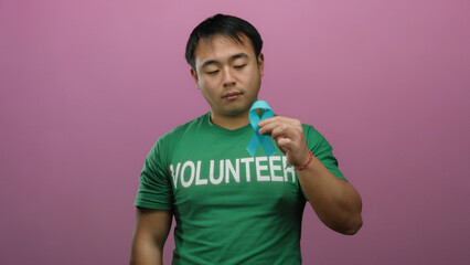 Young chinese man volunteer in green shirt holds blue ribbon against pink background, symbolizing...