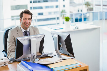 Fototapeta premium Mid adult man wearing suit and tie smiling and sitting at office desk with two monitors