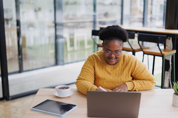 Young black female freelancer smiling while working remotely in a cozy cafe, using a laptop and...