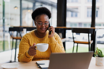 Young African-American businesswoman sipping coffee while engaging in a phone conversation and...