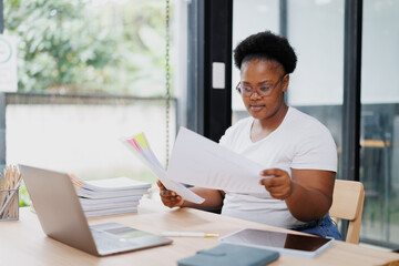 Young office worker is carefully examining a stack of papers at her modern workspace, demonstrating...