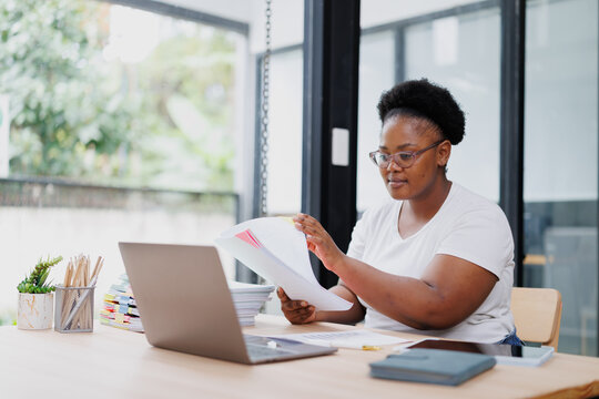 Young black entrepreneur woman reviewing documents and working on laptop at home office, managing her business and finances with concentration and efficiency