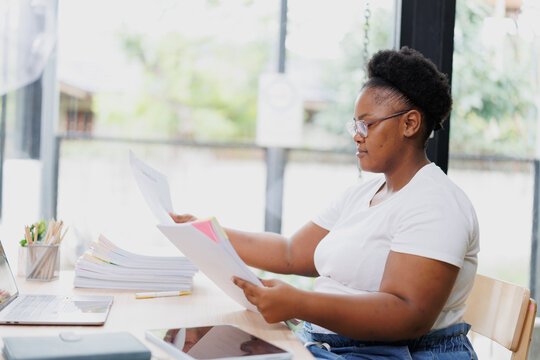 Young african american businesswoman wearing glasses is sitting at her desk and reviewing documents, surrounded by office supplies and technology, in a bright and airy home office setting