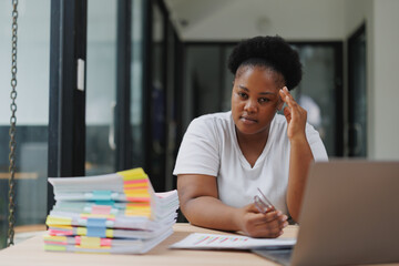 African businesswoman experiencing headache and stress while working on a laptop and documents in a modern office, dealing with chronic pain and seeking medical help