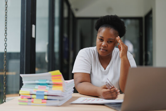 African businesswoman experiencing headache and stress while working on a laptop and documents in a modern office, dealing with chronic pain and seeking medical help