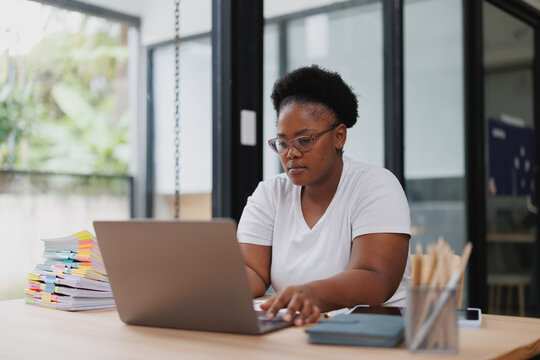 Young black woman running a small business from home, managing her online store and working remotely using her laptop, surrounded by paperwork and office supplies