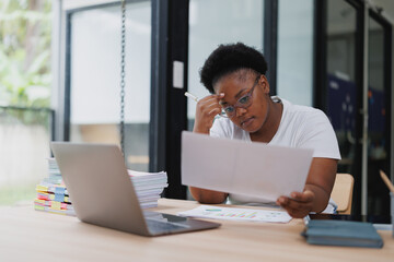 Young african american businesswoman looking concerned while reviewing paperwork and holding head with hand, sitting at desk with laptop and stack of documents in home office