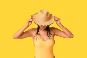 Beautiful young African-American woman in stylish swimsuit and wicker hat on yellow background
