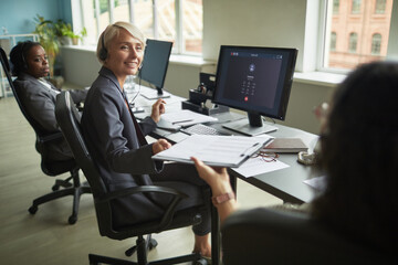 Caucasian middle aged woman wearing headset sitting at desk in modern office, turning to colleague, while Black young adult woman working at computer in background