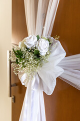A beautiful arrangement of white roses and delicate greenery adorns a wooden door, tied with sheer white fabric. This charming decor creates a welcoming atmosphere at an event.