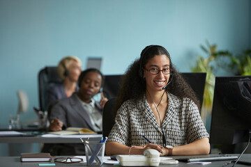 Portrait of young adult woman wearing headset smiling at desk in modern office, sitting in front of computer monitor with colleagues working in background, demonstrating customer support role