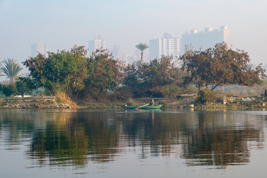 View of a lone figure rowing a small boat on the tranquil river, reflecting the trees and distant buildings under a hazy sky, Zamalek, Cairo Governorate, Egypt.