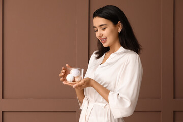 Happy young woman in bathrobe holding bowl with bath bombs on brown background