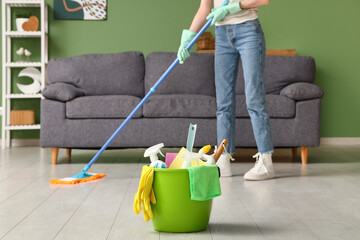 Young woman mopping floor in living room, focus on bucket with cleaning supplies