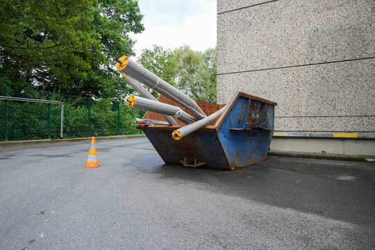 Baucontainer mit Metallteilen &ndash; Industrie-Recycling und Entsorgung
Ein blauer Baucontainer steht neben einem Geb&auml;ude und ist mit gro&szlig;en Metallrohren gef&uuml;llt. Davor ein Verkehrskegel zur Absicherung. S