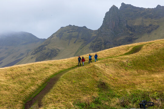 View of hikers traversing a grassy hill under a cloudy sky, with jagged peaks rising in the background in Vik, Iceland, Vik, Iceland.