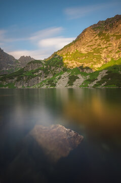 View of a serene lake mirrors the rugged, sunlit mountains, a solitary rock breaking the water's surface in the picturesque landscape, Morskie Oko, Poland.