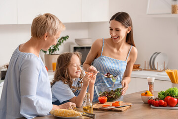 Little girl with her mother and granny eating vegetable salad at table in kitchen