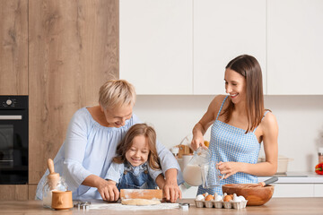 Little girl with her mother and granny rolling out dough at table in kitchen
