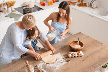 Little girl with her mother and granny rolling out dough at table in kitchen
