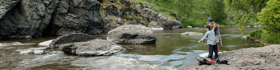 Family with child fishing together on rocky riverbank, standing near flowing water with fishing rods, surrounded by large rocks and lush green trees in outdoor nature setting